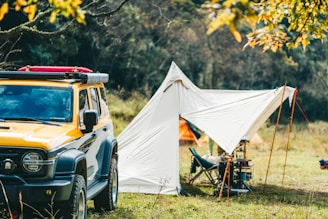 A scene in nature featuring a yellow off-road vehicle parked beside a white tent. The tent is pitched on a grassy field surrounded by trees, with camping equipment visible nearby. The autumn leaves on the trees add vibrant colors to the scene.