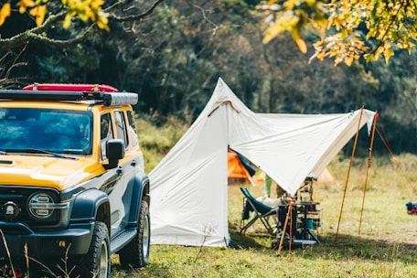A scene in nature featuring a yellow off-road vehicle parked beside a white tent. The tent is pitched on a grassy field surrounded by trees, with camping equipment visible nearby. The autumn leaves on the trees add vibrant colors to the scene.