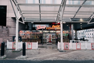 A fast-food restaurant entrance featuring a neon sign with the name Daisy's Express and advertisements for Coca-Cola and hamburgers. The storefront is framed by a white picket fence and a glass canopy overhead. Inside, various signs and a well-stocked beverage cooler are visible. The setting includes a paved sidewalk and nearby urban structures.