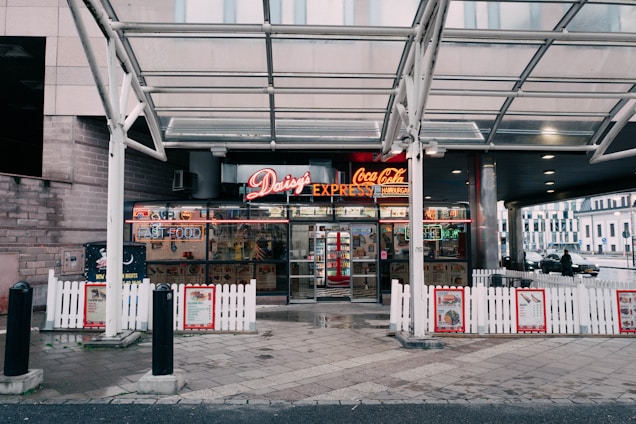 A fast-food restaurant entrance featuring a neon sign with the name Daisy's Express and advertisements for Coca-Cola and hamburgers. The storefront is framed by a white picket fence and a glass canopy overhead. Inside, various signs and a well-stocked beverage cooler are visible. The setting includes a paved sidewalk and nearby urban structures.