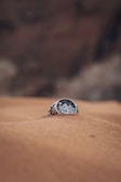 A beautifully crafted silver ring with a detailed engraving of mountains and trees is partially buried in sand. The background is blurred and earthy, adding to the rustic feel of the scene.