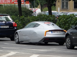 A sleek, futuristic-looking silver car is parked on a street lined with trees and other vehicles. It features aerodynamic design elements such as covered rear wheels and a smooth body. The car is positioned between two darker vehicles on the road.