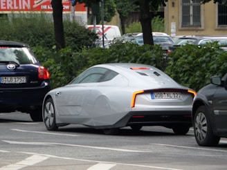 A sleek, futuristic-looking silver car is parked on a street lined with trees and other vehicles. It features aerodynamic design elements such as covered rear wheels and a smooth body. The car is positioned between two darker vehicles on the road.