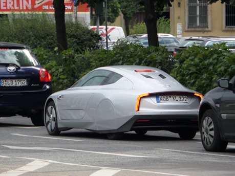 A sleek, futuristic-looking silver car is parked on a street lined with trees and other vehicles. It features aerodynamic design elements such as covered rear wheels and a smooth body. The car is positioned between two darker vehicles on the road.