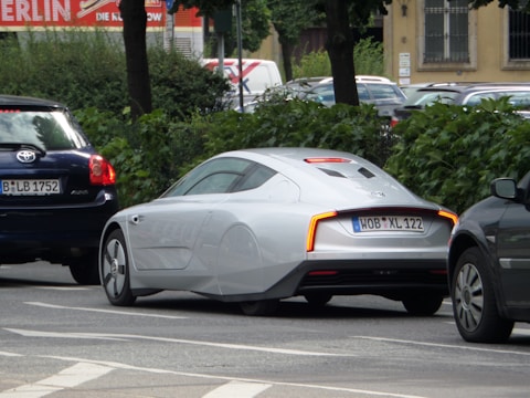 A sleek, futuristic-looking silver car is parked on a street lined with trees and other vehicles. It features aerodynamic design elements such as covered rear wheels and a smooth body. The car is positioned between two darker vehicles on the road.