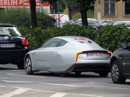 A sleek, futuristic-looking silver car is parked on a street lined with trees and other vehicles. It features aerodynamic design elements such as covered rear wheels and a smooth body. The car is positioned between two darker vehicles on the road.
