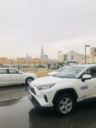 A street scene featuring parked cars, with a prominent white SUV labeled 'URS' in the foreground. The background showcases a cityscape with notable modern architecture, including a distinctive triangular skyscraper. The scene has an overcast sky, with wet pavement indicating recent rain.