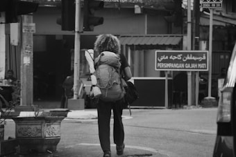 A person with curly hair is walking down a street with a large backpack. The setting appears to be urban with a variety of signs and storefronts. The scene is monochrome, suggesting an overcast day. There are visible traffic lights and a road sign with both Arabic and Roman script.