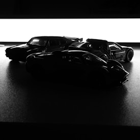 Three model cars are arranged in silhouette against a bright white background, emphasizing their shapes and contours. The lighting creates strong contrasts, highlighting the cars' sleek designs.