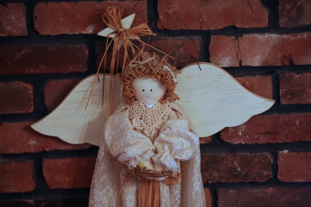 A decorative angel doll with a lace dress and wooden wings, mounted against a red brick wall. The doll has a simple, smiling face and straw-like hair, holding a small basket. A star made of straw-like material is attached to its hair.