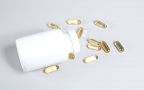 A white plastic bottle lies on its side on a light surface, with several translucent, amber-colored capsules spilling out onto the surface.