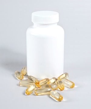 A white plastic bottle with a closed lid is surrounded by several yellowish translucent capsules scattered on a light surface.