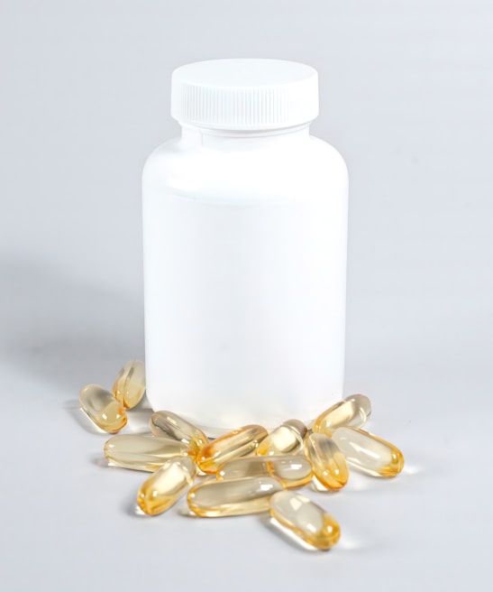 A white plastic bottle with a closed lid is surrounded by several yellowish translucent capsules scattered on a light surface.