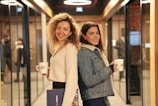 Two women are standing back-to-back in a hallway lined with glass walls and doorways. They are both smiling and holding mugs labeled 'Demo Day'. One woman has curly hair and is wearing a white sweater, holding a folder. The other woman has long dark hair and is wearing a denim jacket with a shearling collar.