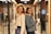 Two women are standing back-to-back in a hallway lined with glass walls and doorways. They are both smiling and holding mugs labeled 'Demo Day'. One woman has curly hair and is wearing a white sweater, holding a folder. The other woman has long dark hair and is wearing a denim jacket with a shearling collar.