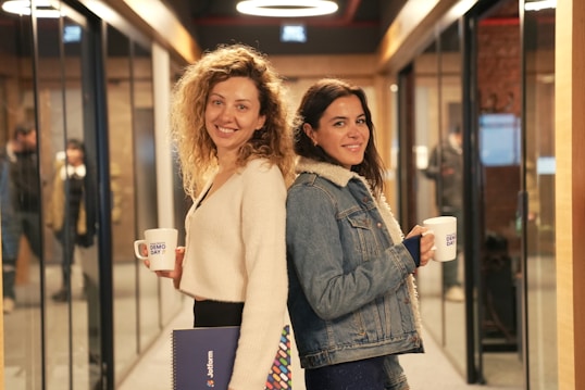 Two women are standing back-to-back in a hallway lined with glass walls and doorways. They are both smiling and holding mugs labeled 'Demo Day'. One woman has curly hair and is wearing a white sweater, holding a folder. The other woman has long dark hair and is wearing a denim jacket with a shearling collar.