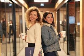Two women are standing back-to-back in a hallway lined with glass walls and doorways. They are both smiling and holding mugs labeled 'Demo Day'. One woman has curly hair and is wearing a white sweater, holding a folder. The other woman has long dark hair and is wearing a denim jacket with a shearling collar.
