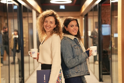 Two women are standing back-to-back in a hallway lined with glass walls and doorways. They are both smiling and holding mugs labeled 'Demo Day'. One woman has curly hair and is wearing a white sweater, holding a folder. The other woman has long dark hair and is wearing a denim jacket with a shearling collar.