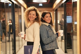Two women are standing back-to-back in a hallway lined with glass walls and doorways. They are both smiling and holding mugs labeled 'Demo Day'. One woman has curly hair and is wearing a white sweater, holding a folder. The other woman has long dark hair and is wearing a denim jacket with a shearling collar.