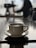 A steaming cup of coffee sits on a saucer, placed on a reflective surface. In the background, there is a blurred silhouette of a person, possibly a barista, adjusting a coffee machine. The lighting is soft, with a moody and atmospheric feel.