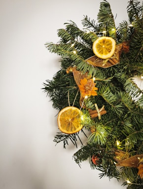 A festive wreath made of evergreen branches adorned with dried orange slices, golden ribbons, cinnamon sticks, and small white lights. The wreath is hanging against a plain, light-colored background, creating a simple yet elegant holiday decoration.