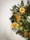 A festive wreath made of evergreen branches adorned with dried orange slices, golden ribbons, cinnamon sticks, and small white lights. The wreath is hanging against a plain, light-colored background, creating a simple yet elegant holiday decoration.