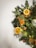 A festive wreath made of evergreen branches adorned with dried orange slices, golden ribbons, cinnamon sticks, and small white lights. The wreath is hanging against a plain, light-colored background, creating a simple yet elegant holiday decoration.