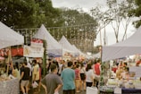 A bustling outdoor market with various stalls under white tents, illuminated by string lights. People are walking around, exploring the goods and food items on display. The atmosphere is lively with a mix of vendors and visitors.