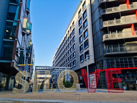 Large metallic letters spell out 'SLOVE' and are set against a modern urban backdrop with multi-story buildings and clear blue skies.