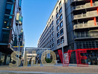 Large metallic letters spell out 'SLOVE' and are set against a modern urban backdrop with multi-story buildings and clear blue skies.