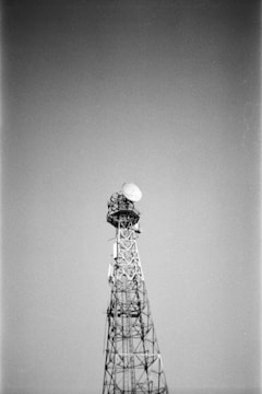 A tall metal communication tower stands against a clear sky. The structure is composed of a lattice framework with a large satellite dish mounted near the top.