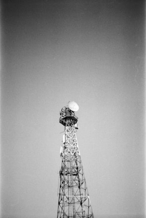 A tall metal communication tower stands against a clear sky. The structure is composed of a lattice framework with a large satellite dish mounted near the top.