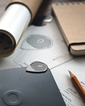A close-up view of a workspace with a notebook, a pencil, a rolled paper tube, and a sheet of paper with detailed design sketches. A textured, metal guitar pick or similar object rests on a dark surface, possibly a tablet or notebook. The setting is organized and focused on design or planning work.