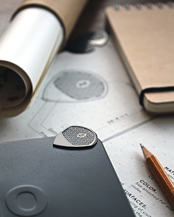 A close-up view of a workspace with a notebook, a pencil, a rolled paper tube, and a sheet of paper with detailed design sketches. A textured, metal guitar pick or similar object rests on a dark surface, possibly a tablet or notebook. The setting is organized and focused on design or planning work.