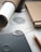 A close-up view of a workspace with a notebook, a pencil, a rolled paper tube, and a sheet of paper with detailed design sketches. A textured, metal guitar pick or similar object rests on a dark surface, possibly a tablet or notebook. The setting is organized and focused on design or planning work.