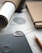 A close-up view of a workspace with a notebook, a pencil, a rolled paper tube, and a sheet of paper with detailed design sketches. A textured, metal guitar pick or similar object rests on a dark surface, possibly a tablet or notebook. The setting is organized and focused on design or planning work.