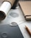A close-up view of a workspace with a notebook, a pencil, a rolled paper tube, and a sheet of paper with detailed design sketches. A textured, metal guitar pick or similar object rests on a dark surface, possibly a tablet or notebook. The setting is organized and focused on design or planning work.