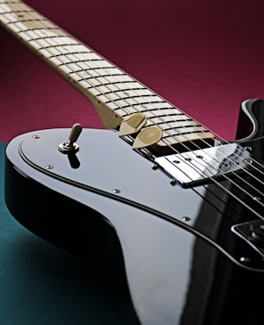 A close-up view of a glossy black electric guitar, focusing on its body and neck. The strings, pickups, and control knobs are clearly visible, while two guitar picks rest on the strings. The background is a blend of deep burgundy and dark teal, complementing the sleek surface of the instrument.