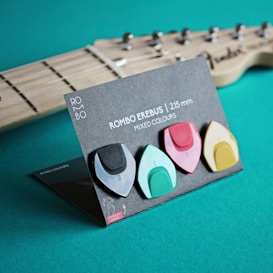 Four guitar picks, each in different colors, are displayed on a cardboard holder. They are arranged neatly in a row with the product details 'ROMBO EREBUS | 2.15 mm' written above them. A guitar neck is partially visible in the background.