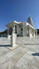 A grand white marble temple with intricate carvings stands under a clear blue sky. The temple is elevated with stairs leading up to its entrance. Columns support a large, ornate roof, and the structure features a prominent tower or spire. A sign indicating 'offerings' is placed on a pedestal in the foreground. Few people are visible, providing a sense of scale to the large, open courtyard.