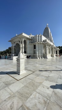 A grand white marble temple with intricate carvings stands under a clear blue sky. The temple is elevated with stairs leading up to its entrance. Columns support a large, ornate roof, and the structure features a prominent tower or spire. A sign indicating 'offerings' is placed on a pedestal in the foreground. Few people are visible, providing a sense of scale to the large, open courtyard.
