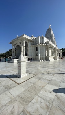 A grand white marble temple with intricate carvings stands under a clear blue sky. The temple is elevated with stairs leading up to its entrance. Columns support a large, ornate roof, and the structure features a prominent tower or spire. A sign indicating 'offerings' is placed on a pedestal in the foreground. Few people are visible, providing a sense of scale to the large, open courtyard.