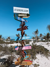 A colorful signpost with multiple directional arrows stands on a sandy beach surrounded by palm trees. Each arrow points towards different cities, including Brasilia, Ottawa, Buenos Aires, Holbox, Isla Mujeres, and Bogota, with distances listed in kilometers and miles. The sky is clear and blue, creating a tropical and inviting atmosphere.
