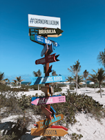 A colorful signpost with multiple directional arrows stands on a sandy beach surrounded by palm trees. Each arrow points towards different cities, including Brasilia, Ottawa, Buenos Aires, Holbox, Isla Mujeres, and Bogota, with distances listed in kilometers and miles. The sky is clear and blue, creating a tropical and inviting atmosphere.