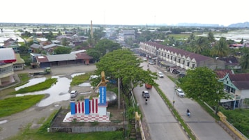 Aerial view of a small town featuring a main road, various buildings, and green spaces. There is a monument or statue with patriotic colors in the foreground. Cars and motorcycles are seen on the road, with many parked near buildings. Trees are scattered around the area, and there are fields in the background.