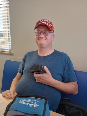 A man wearing glasses and a red cap with political branding, holding a smartphone. He is seated at a table with a blue and gray fabric item in front of him. The background includes a partial view of a window with blinds.