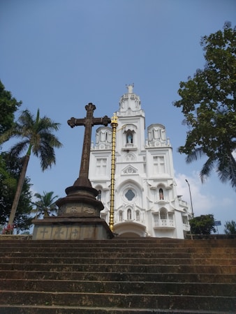 A grand white church with intricate architectural details stands prominently against a blue sky. A large, ornate cross and a gold-colored pillar are positioned in front of the church, surrounded by lush green trees.
