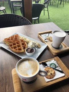 A wooden table is set with two trays, each containing a steaming cup of coffee and a cookie on a napkin. In the middle, a white plate holds waffles, banana slices, whipped cream, and a small bowl of chocolate spread. The background features wicker chairs and tables on a green artificial turf.
