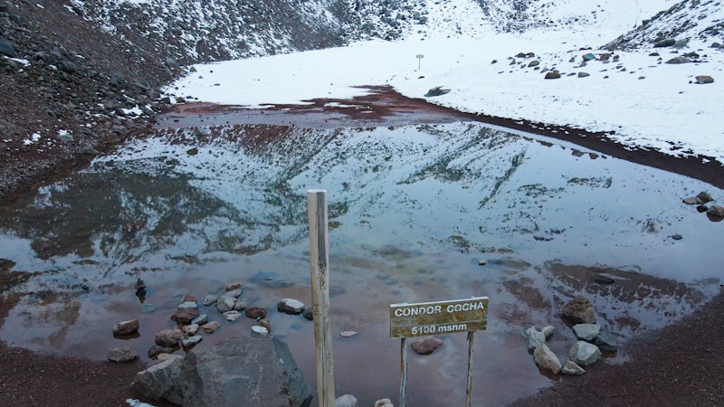 A partially frozen mountain lake surrounded by snow-covered slopes and rocks. The water reflects the snowy landscape and nearby hills. A wooden signpost indicates the name 'CONDOR COCHA' and an elevation of 5100 meters above sea level.