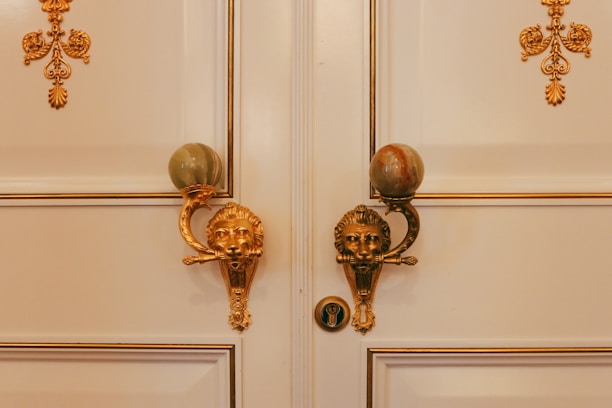 An ornate double door with elegant golden lion head door knockers, each holding a large spherical stone. Intricate gold decorations adorn the white door panels, adding a luxurious and decorative touch.